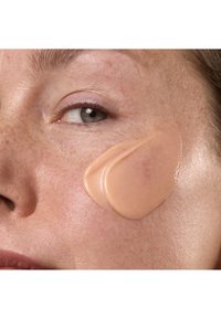 Crema beige applicata sulla pelle con texture visibile; primo piano di un volto che mostra lentiggini, pelle liscia e un trucco naturale.