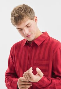 Camisa roja de botones, con rayas verticales, mangas largas y un bolsillo en el pecho. La tela parece suave y texturada. Modelo ajustándose el puño.