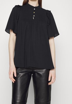 Woman wearing a loose black blouse with ruffled shoulders and three small buttons, paired with black leather pants.