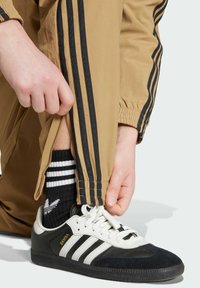 Black and white sneakers with suede and leather accents, paired with tan track pants featuring black stripes and a zippered ankle.
