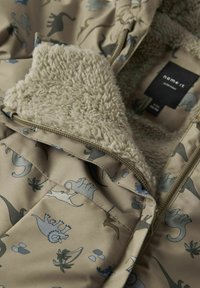 Veste imprimée de dinosaures beige avec une doublure intérieure douce et pelucheuse et une fermeture éclair dorée. Le tissu est lisse avec une texture matelassée.