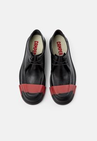 Schwarze Lederschuhe mit einer abgerundeten Spitze und einem markanten roten Gummi-Band-Akzent über der Zehenpartie. Mit einer Schnürung und einem neutralen Innenraum.