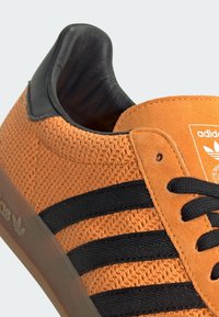 Narančaste nubuk tenisice s crnim tekstilnim trakama, crnim petnim dijelom i crnim vezicama. Imaju teksturiranu površinu i oznaku s logotipom na jeziku.