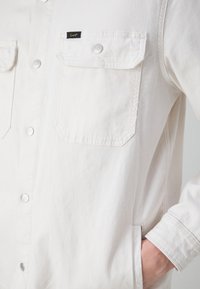 Camisa de mezclilla blanca con cierres plateados, dos bolsillos en el pecho, solapas de bolsillo en ángulo y un pequeño logo negro "Lee" en el bolsillo izquierdo.