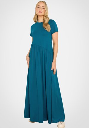 Mujer con pelo largo y rubio, vestida con un vestido maxi de manga corta color verde azulado y zapatillas blancas, de pie sobre un fondo claro y liso.
