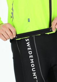 Neon gul cykeljakke med sorte accenter og et svensk flag, parret med sorte bukser med "SWEDEMOUNT" i hvidt bogstaver.