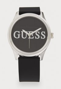 Reloj de goma negra con caja de metal de tono plateado, esfera redonda y audaz logotipo "GUESS". Presenta un dial oscuro texturizado y manecillas plateadas.