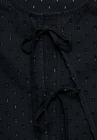 Crni materijal s malim, sjajnim ukrasima. Ima okrugli izrez, vezanje sprijeda i teksturiranu površinu.