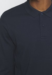 Mörkblå långärmad polo med knäppning, i bomullsmaterial och ribbad krage. Har diskret sömnad och inga synliga mönster.