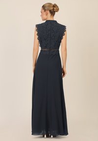 Abito in pizzo blu navy con maniche a coste, dotato di un corpo in pizzo aderente e una gonna fluida. Chiusura posteriore con dettaglio zipper.