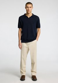 Dark navy knitted polo shirt with short sleeves, paired with light beige trousers and brown moccasin-style shoes. Simple design and texture.