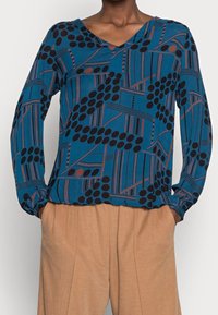 Langärmlige, türkisfarbene Bluse mit einem geometrischen Muster aus schwarzen Kreisen und braunen Linien. Der Stoff wirkt weich und hat ein V-Ausschnitt-Design.