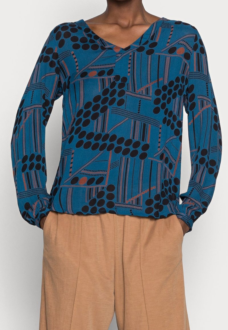 Langärmlige, türkisfarbene Bluse mit einem geometrischen Muster aus schwarzen Kreisen und braunen Linien. Der Stoff wirkt weich und hat ein V-Ausschnitt-Design.