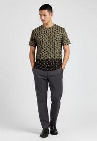 Krátký rukáv tričko v olivově zelené s černým geometrickým vzorem, kombinováno se šedými kalhotami a černými botami. Hladký povrch tkaniny.