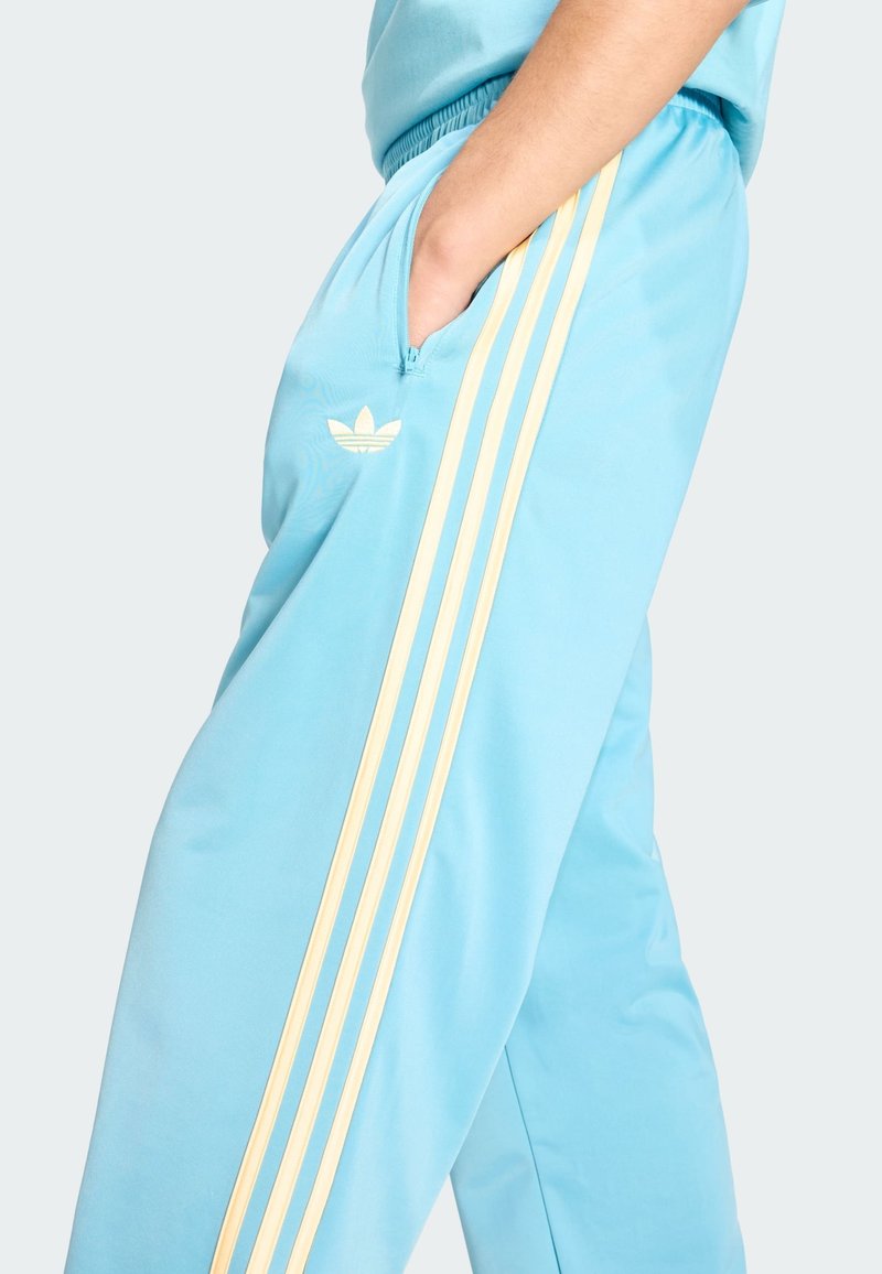 Persona che indossa pantaloni della tuta Adidas azzurri con tre strisce gialle e logo, mano nella tasca con cerniera destra, su sfondo semplice.