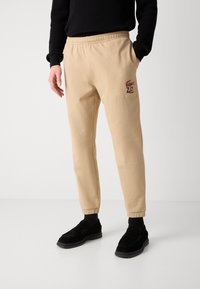 Béžové tepláky s pohodlným strihom, elastickým pásom a manžetami na členkoch. Na ľavej strane sa nachádza červené vyšívané logo Lacoste.