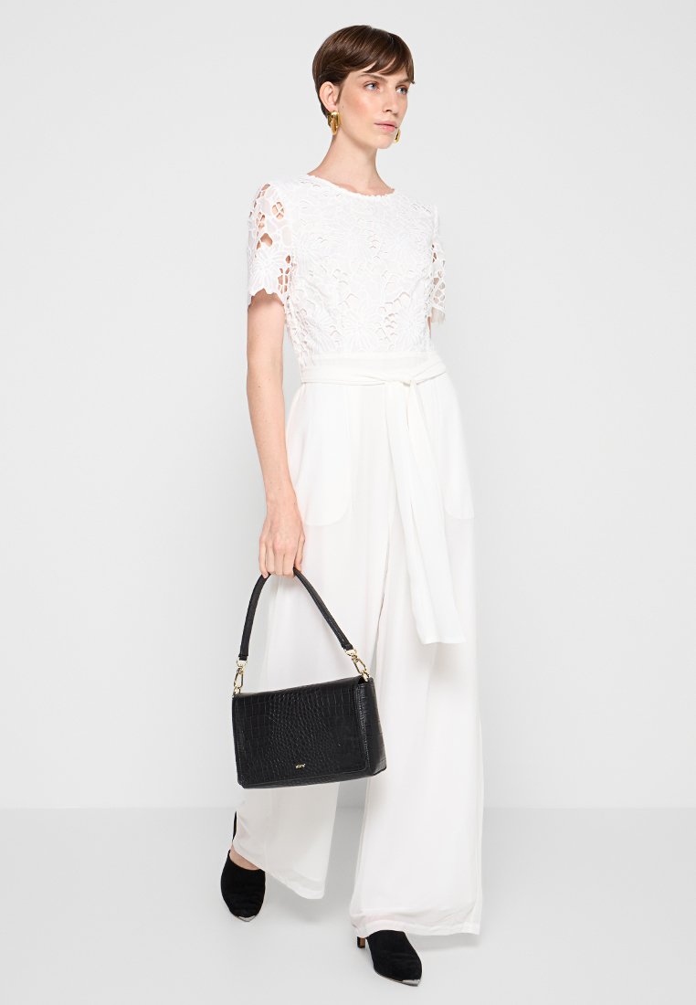 Tuta bianca con dettagli in pizzo, gambe larghe e vita legata. Borsa nera con texture in croco e manici corti. Scarpe nere.