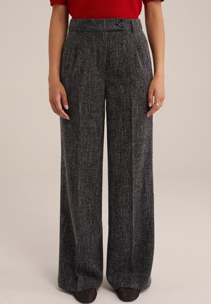 Vrouw draagt high-waist wijde donkere grijze textuurbroek met knoopsluiting en zwarte mesh schoenen, staand tegen een effen achtergrond.