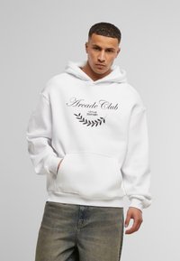 Felpa bianca con cappuccio, dotata di tasca a marsupio frontale e testo nero che dice "Arcade Club" con un design a forma di foglia, abbinata a jeans chiari.