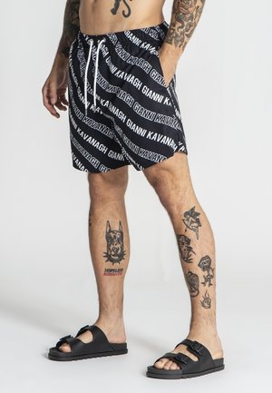 Uomo che indossa pantaloncini Gianni Kavanagh bianco e nero, sandali neri con fibbia, con tatuaggi su gambe e braccia, in piedi contro uno sfondo uniforme.