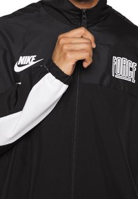 Black and white Nike jacket with "FORCE" text, zipped halfway, hand holding zipper near collar.