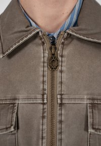 Nærbillede af en delvist lynet brun jakke med en dekorativ messing lynlåstrækker over kraven på en blåstripet skjorte.