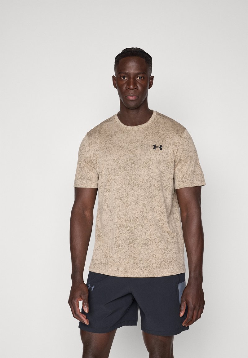 Hombre de pie con una camisa de manga corta texturizada beige y pantalones cortos deportivos negros sobre un fondo claro y liso.