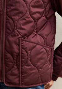 Burgundy quiltad jacka med en strukturerad vågmönster, dragkedjestängning och en sidoficka med matchande kant. Mjuk, lättviktig material.