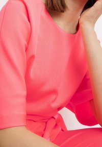 Ljusrosa kortärmad topp i mjukt material, med rund halsringning och en sidoknytning vid midjan.