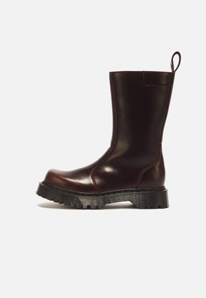 Dunkelbrauner Lederstiefel bis zur Mitte der Wade mit dicker schwarzer profilierter Sohle und Schlaufe hinten vor weißem Hintergrund.