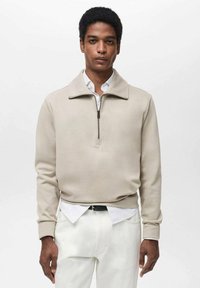 Pull-over beige con zip e colletto alto, realizzato in materiale morbido. Indossato su una camicia bianca con bottoni, abbinato a pantaloni bianchi.