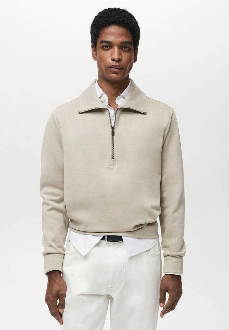 Pull-over beige con zip e colletto alto, realizzato in materiale morbido. Indossato su una camicia bianca con bottoni, abbinato a pantaloni bianchi.