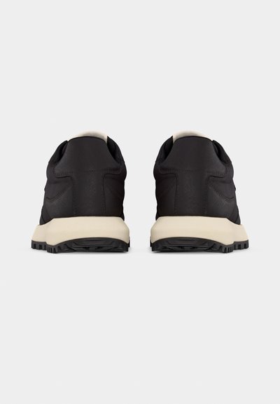 Ζευγάρι μαύρων αθλητικών παπουτσιών με μπεζ σόλες, που φαίνεται από πίσω, αναδεικνύοντας την υφασμένη φτέρνα και το πρότυπο πέλματος.