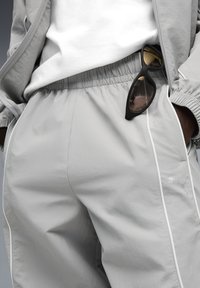 Persoon in lichtgrijze Puma trainingsbroek en -jack met een wit shirt, zwarte zonnebril aan de tailleband van de broek.