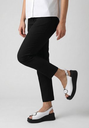 Kobieta ubrana w czarne spodnie i białe, na platformie, sandały z odkrytymi palcami i srebrną klamrą, stojąca z jedną nogą skrzyżowaną na drugą.