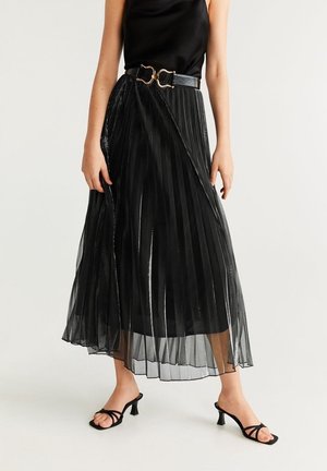 Frau trägt schwarzen ärmellosen Oberteil, schwarzen plissierten transparenten Rock mit Gürtel und schwarze offene Sandaletten mit Absatz, steht auf weißem Boden.