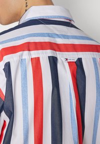 Camicia a righe nei colori bianco, rosso, blu e denim; presenta un colletto classico e un'etichetta con il logo sul retro. Tessuto di cotone morbido.