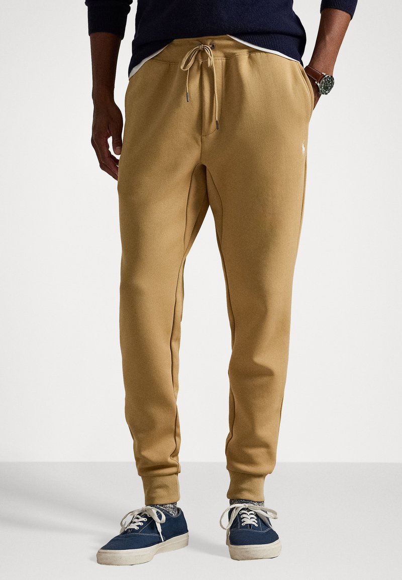 Beige sweatpants with a relaxed fit, featuring drawstring cuffs, side pockets, and a small logo on the thigh. Soft textured material.