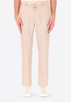 Pantaloni in lino beige con coulisse in vita, indossati con scarpe da ginnastica bianche, mani lungo i fianchi, su uno sfondo chiaro e uniforme.