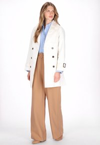 Cremefarvet dobbeltradet trenchcoat med store brune knapper, kombineret med en lyseblå skjorte og tanfarvede viddebukser. Blød tekstur generelt.