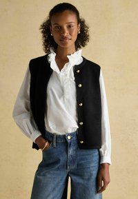 Must must vest kuldnuppudega, mille alla on kantud valge volangiga särk ja sinised teksapüksid. Omab kohandatud lõiget ja tekstuurset kangast.