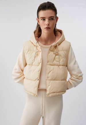 Mlada žena koja nosi bež pamučni prsluk bez rukava s kapuljačom preko kremaste majice i pripadajućih hlača, stoji s rukama iza leđa.