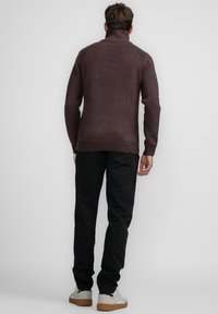Suéter de cuello alto marrón con puños y dobladillo acanalados, combinado con pantalones negros y zapatillas blancas, visto desde la parte posterior.