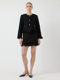 Schwarze Texturjacke mit Rüschensaum und -manschetten, akzentuiert mit silbernen Knöpfen. Kombiniert mit einem passenden schwarzen Rüschensock und schwarzen Absätzen.