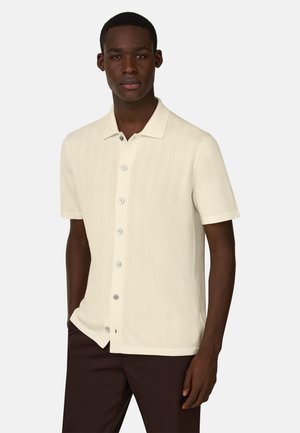Mann trägt ein cremefarbenes, kurzärmeliges Hemd mit Knöpfen und Kragen, kombiniert mit dunkelbraunen Hosen, stehend vor einem einfarbigen Hintergrund.