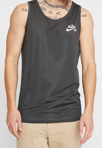 Débardeur en maille noir avec un col rond, présentant de petites perforations et un logo Nike SB blanc sur la poitrine.