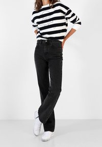 Personne portant un jean noir droit, des baskets blanches et une chemise à manches longues noir et blanche rayée avec un motif de crâne.