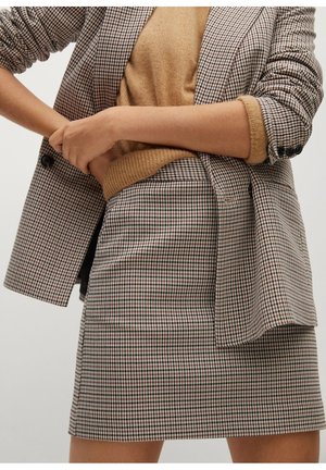 Frau trägt einen karierten Blazer und passenden Rock mit einem camelfarbenen Pullover mit hochgekrempelten Ärmeln, Hände vor der Taille verschränkt.