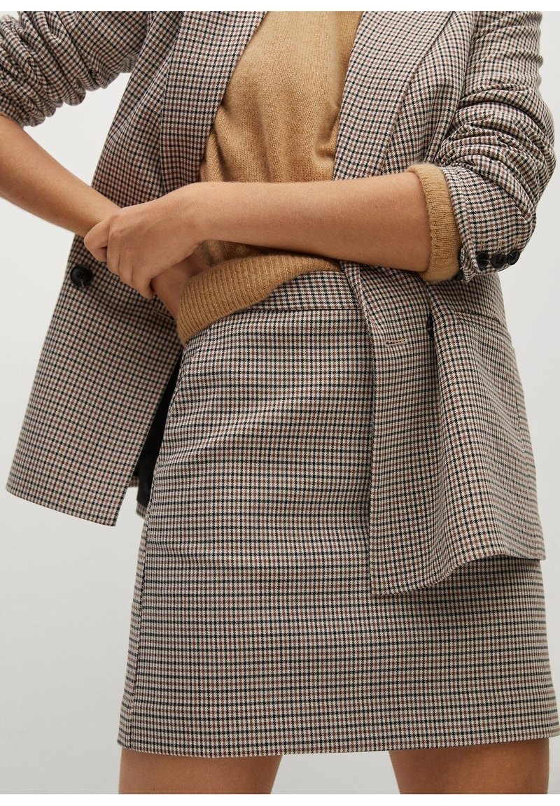 Femme portant un blazer à carreaux et une jupe assortie avec un pull couleur chameau aux manches retroussées, mains croisées à la taille.