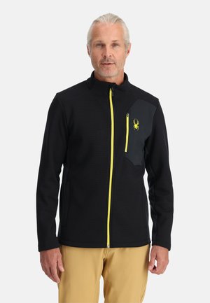 Veste en polaire noire avec fermeture éclair et accents jaunes, présentant un design texturé et une poche noire contrastante avec un logo sur la poitrine.